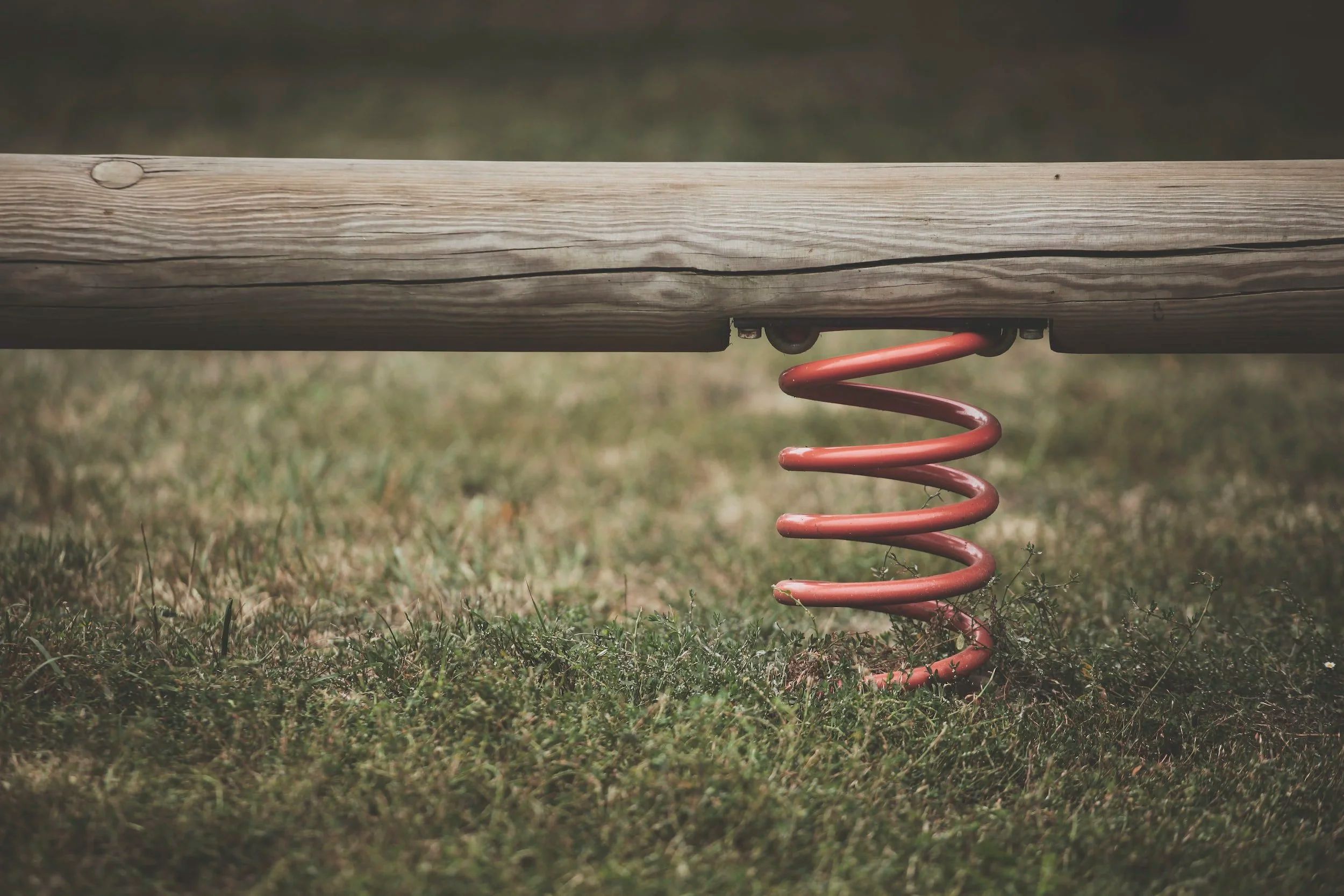 A red coil spring under a wooden beam on a playground, in green grass.