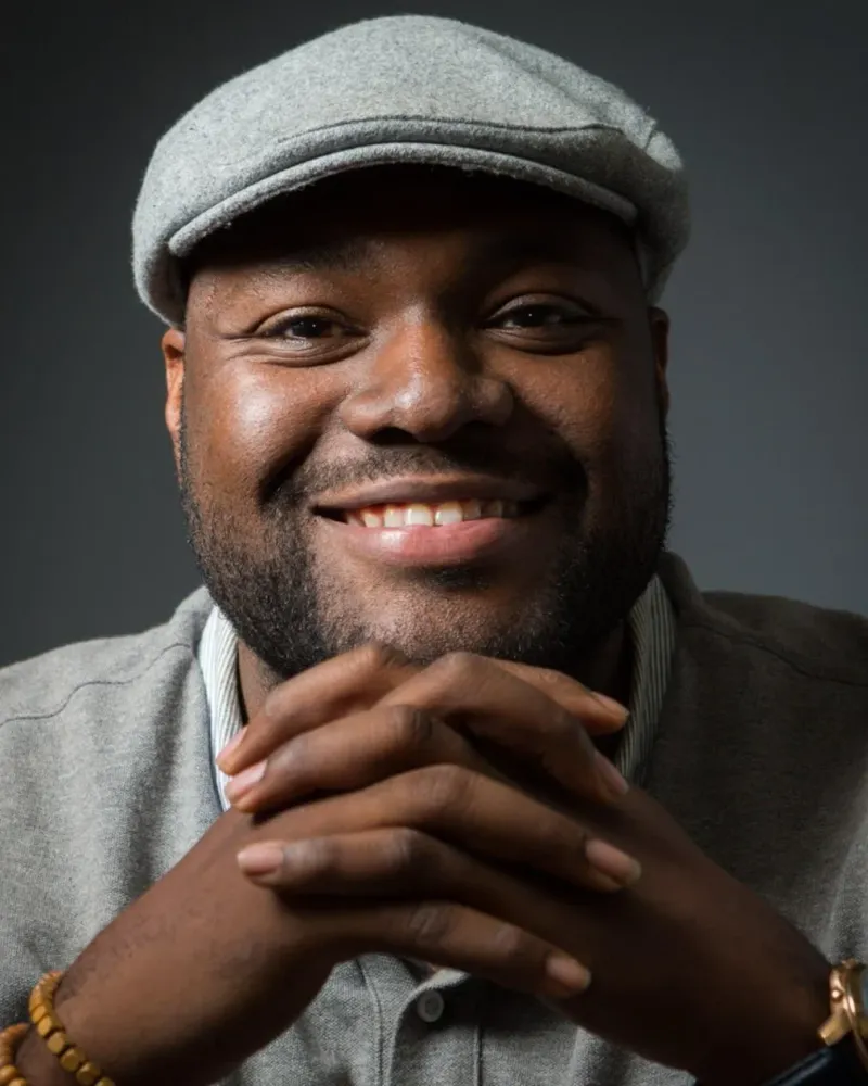 Timothy Ojetunde smiling with his hands clasped, wearing a grey flat cap.