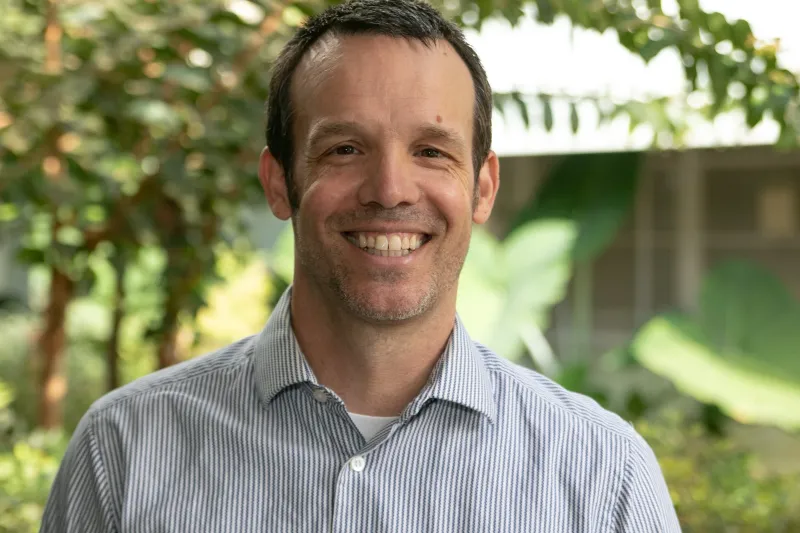Tobin Paap smiling outdoors in a striped shirt, with greenery behind him.